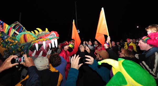 A crowd dressed in winter clothes hold up cameras and crowd around a large dragon head. A man dressed in red, waving an orange flag leads it through the crowd.