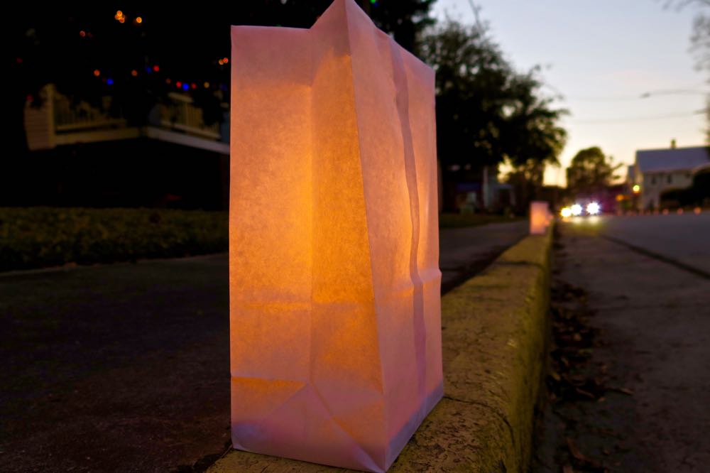 A white, wax bag sits on a street curb. A candle glows inside it.