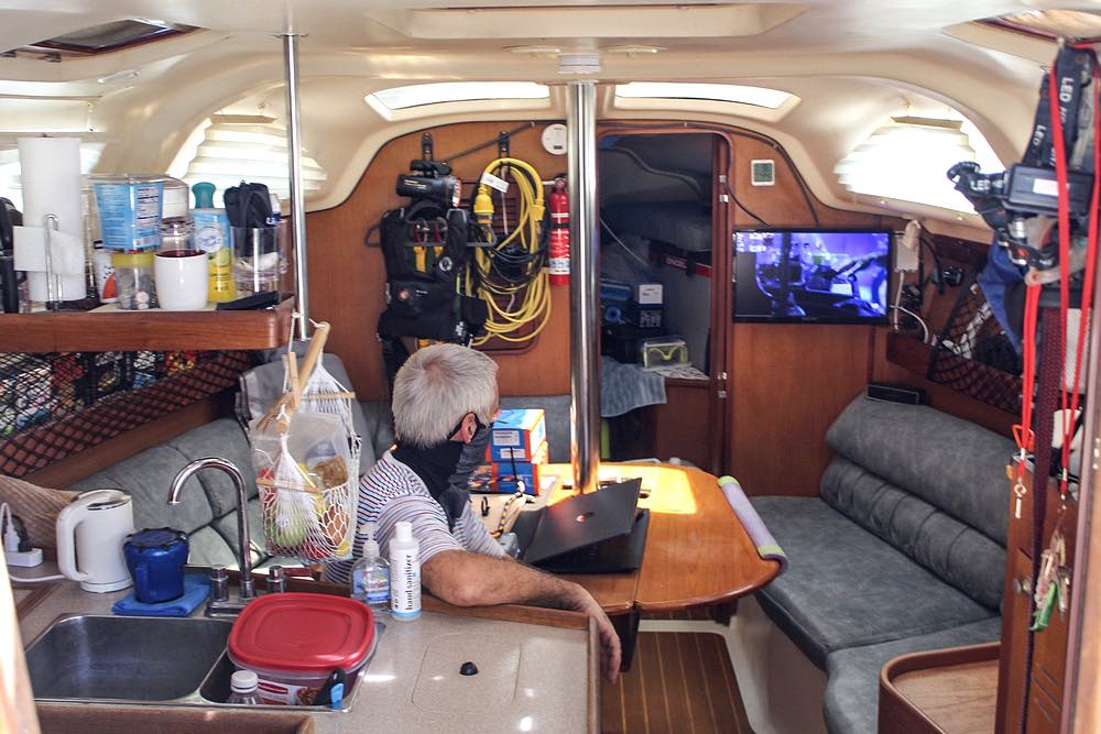 Inside Luna Sea, Mark sits at a table and watches a TV mounted on the wall. Gear hangs from the walls and nets. More gear can be seen through a cabin door.