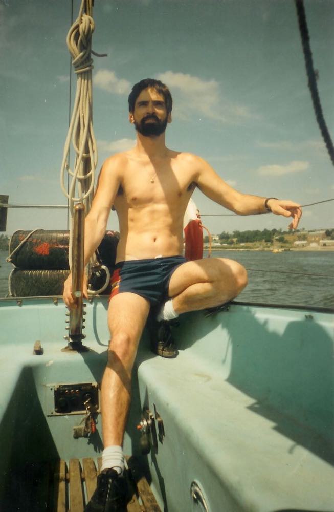 A older photo, probably from the 80s, shows a young man with a beard, wearing socks and tennis shoes, and a pair of dark color shorts at the tiller of a sailboat. The boat's cockpit is light blue and the day is bright.