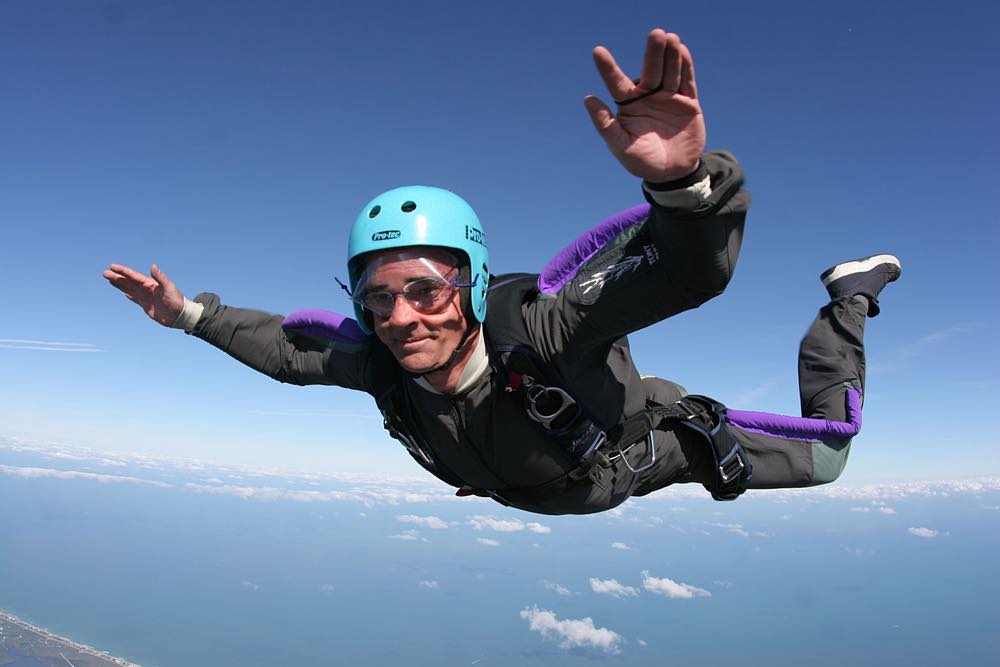 Mark wears a blue helmet and black and purple skydiving suit as he is suspended in midair. His parachute is not yet deployed.
