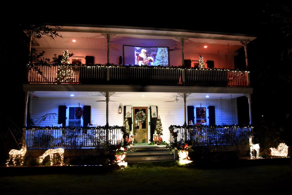 A two story house with red and blue lights, nut crackers, lit deer, and a movie of Santa on the second floor balcony