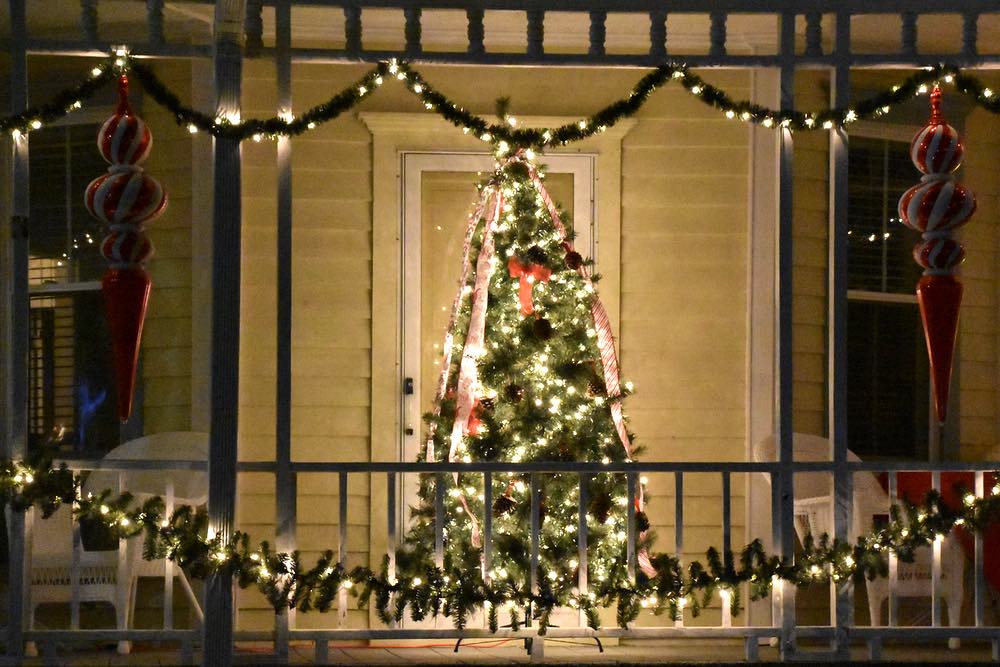 A lit tree sits on a porch decoratedw ith lights and oversized red and white ornaments'