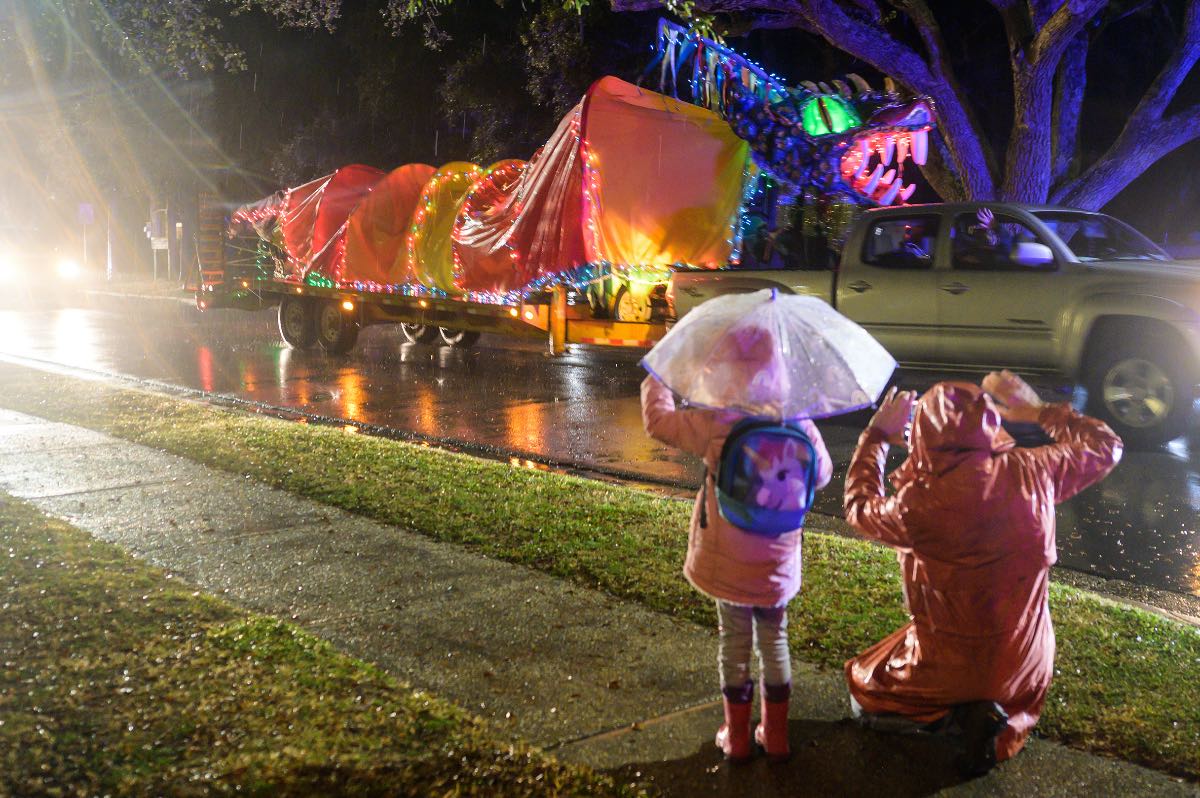 A little girl in a pink raincoat and unicorn backpack holds a clear umbrella. Her grandmother kneels next to her. It's raining and they're waving to a lighted dragon being towed down the street on a trailer.