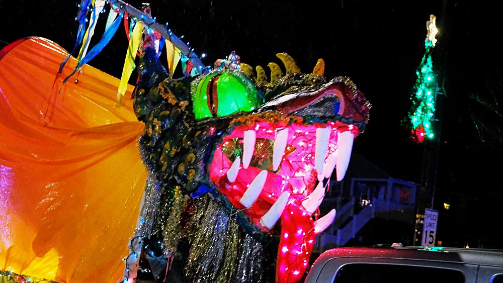 Close up of the dragon head: bright green eyes, pink mouth, white horns with blue and yellow streamers wrapped in blue lights, and an orange body and gold and silver tassel beard.