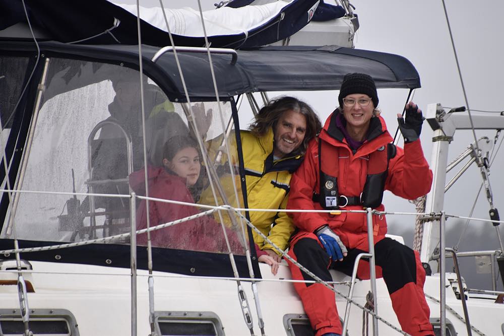 A woman in red foul weather gear waves as a man and young girl look out from the cockpit and smile.