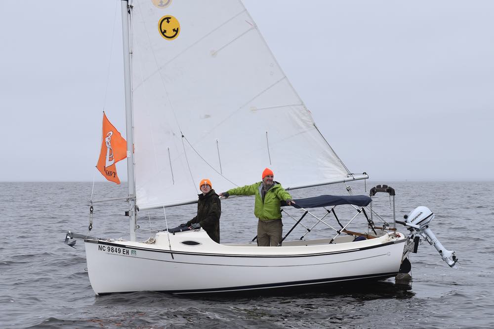 A young man and older man, both wearing bright orange caps, sail by in a small, single masted sailboat. 