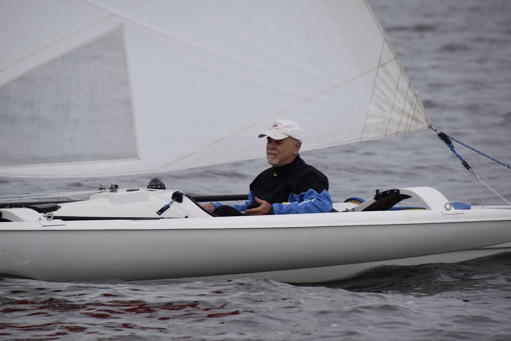 A man in a black and blue foul weather jacket sails a small, white trimaran.