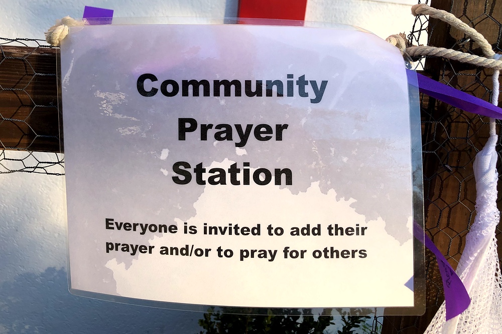 A laminated sign hangs from chicken wire wrapped around a wooden cross stating 'Community Prayer Station.'