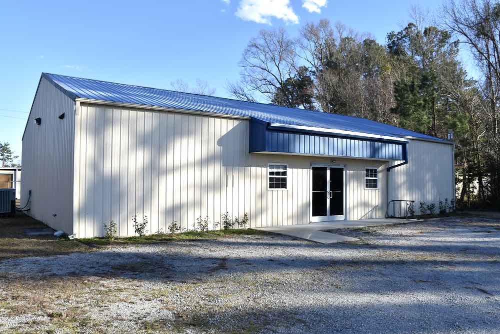 Exterior view of a metal building with double entry doors and a small window on either side of the entry way. A blue awning stretches over the windows and doors. The parking lot is gravel and tall oak and pine trees grow on the building's right side.