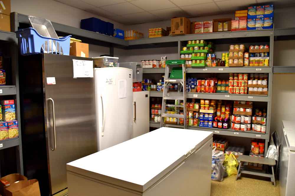 Inside the food pantry are three upright refrigerators: large, medium, and small. One deep freezer sits in the middle of the room, another against the wall. Shelves with boxes of canned goods line the wall. There isn't much space to move around in the pantry.