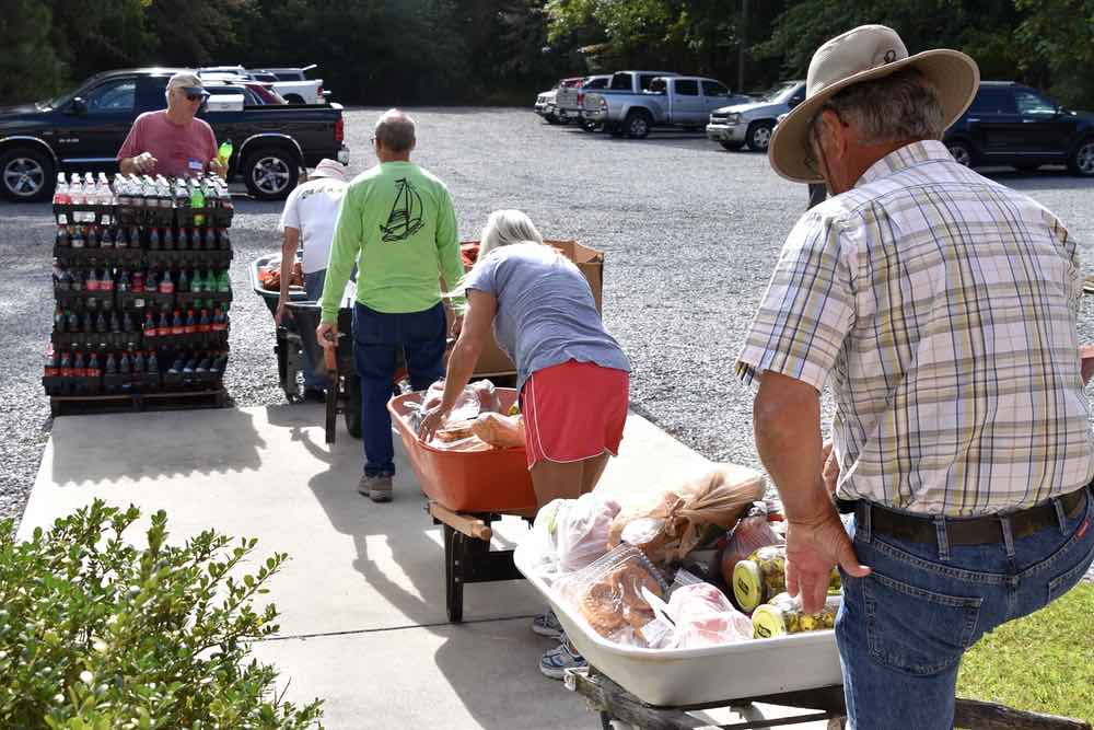 Four volunteers, older men and women, line up in the parking lot with wheelbarrows full of food. Another volunteer stands near a pallet full of soda, waiting to unload these to the waiting vehicles.