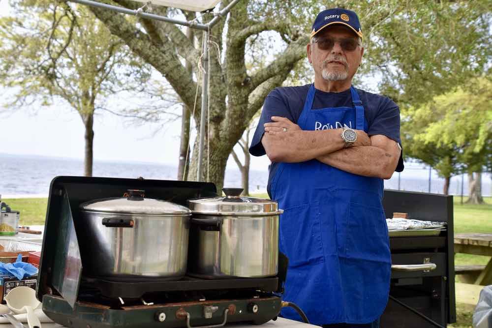 Two large silver cook pots sit on a camp stove. A bearded man wearing a blue apron and hat that both say 'Rotary' stands near by with his arms crossed. 
