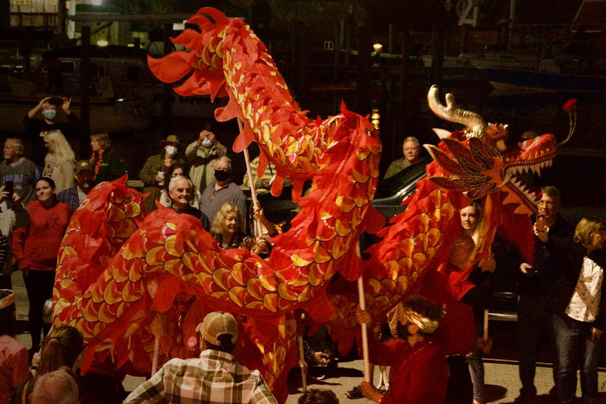 Nighttime on a crowded street over looking dark water in the harbor. The red, yellow, and gold dragon winds around itself above the crowd.
