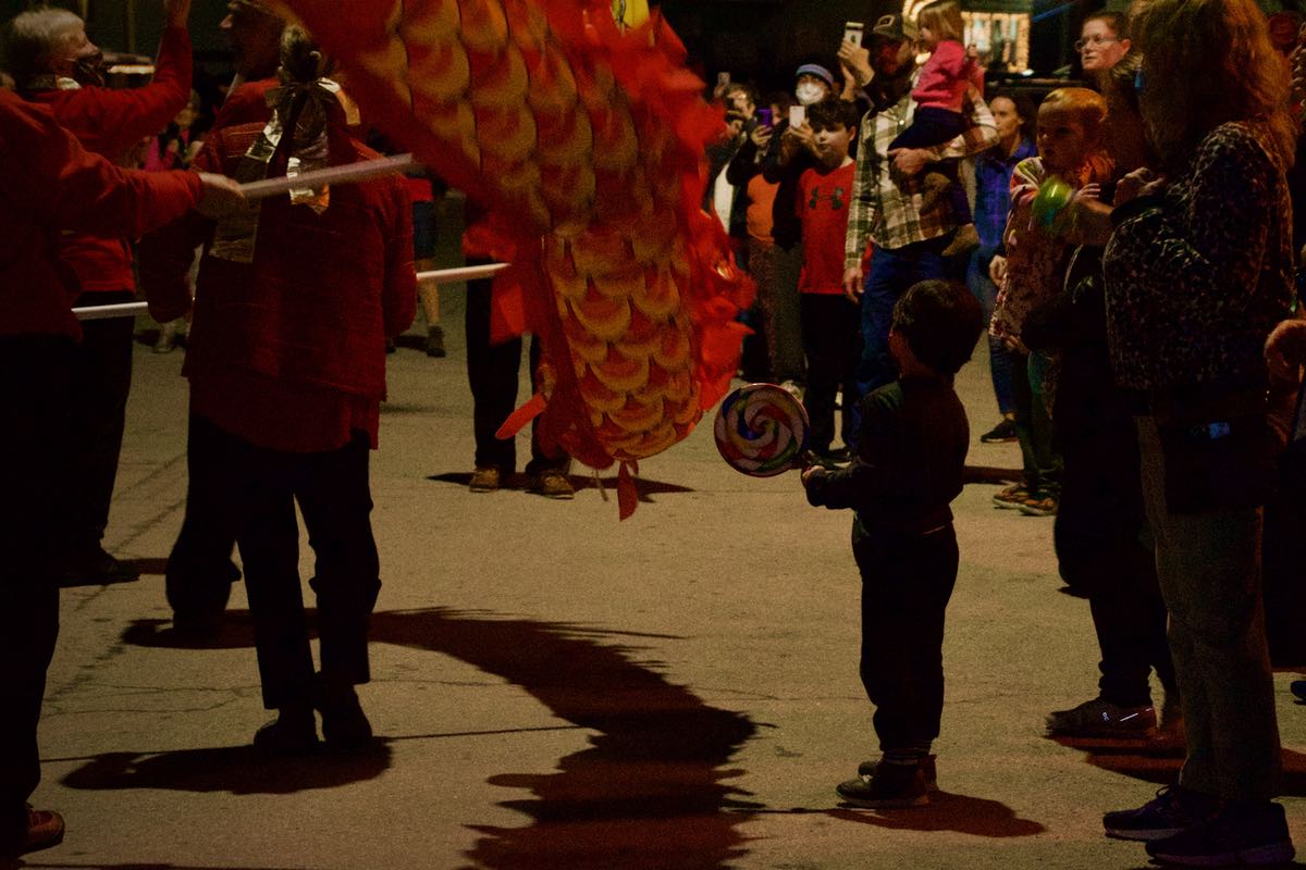 A small boy with a large lollipop stands in the streetlight while the body of the red, yellow, and gold dragon passes next to him. 