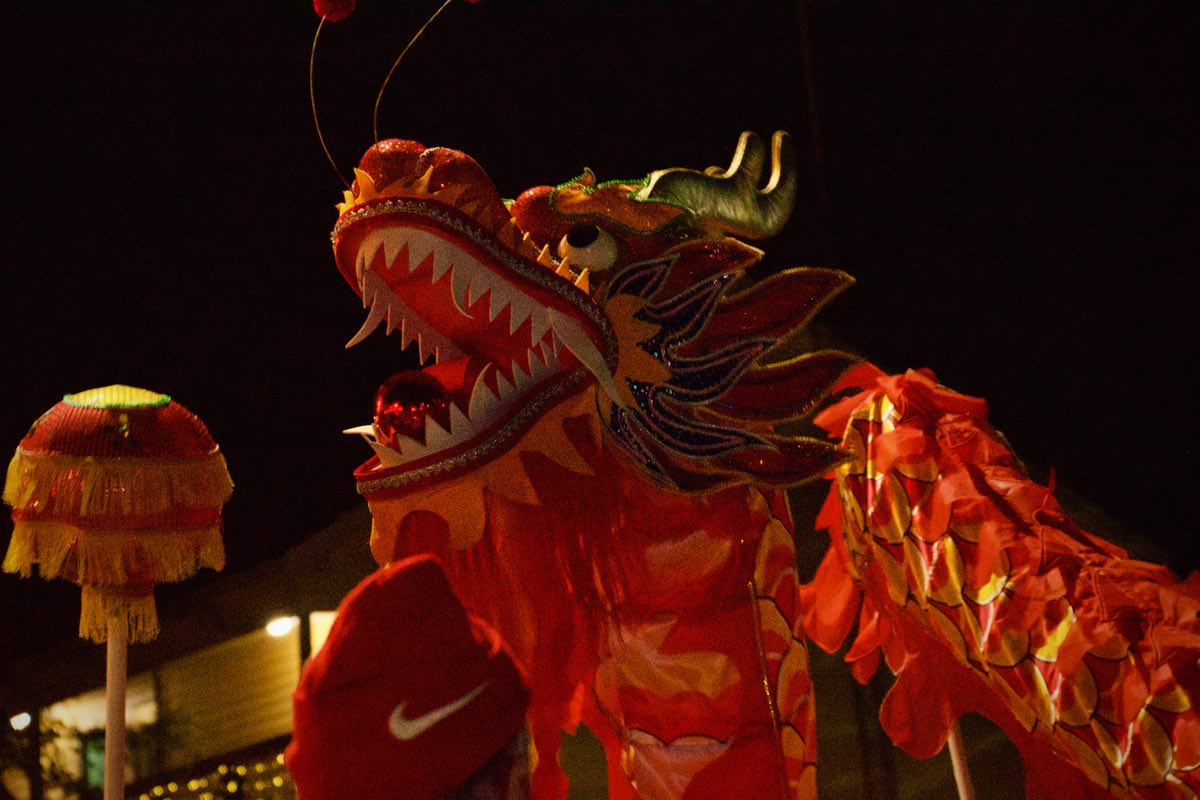 Nighttime. A red, yellow, and gold snake-like dragon head is above the crowd. A large gold colored ball with red fringe is held near its head.
