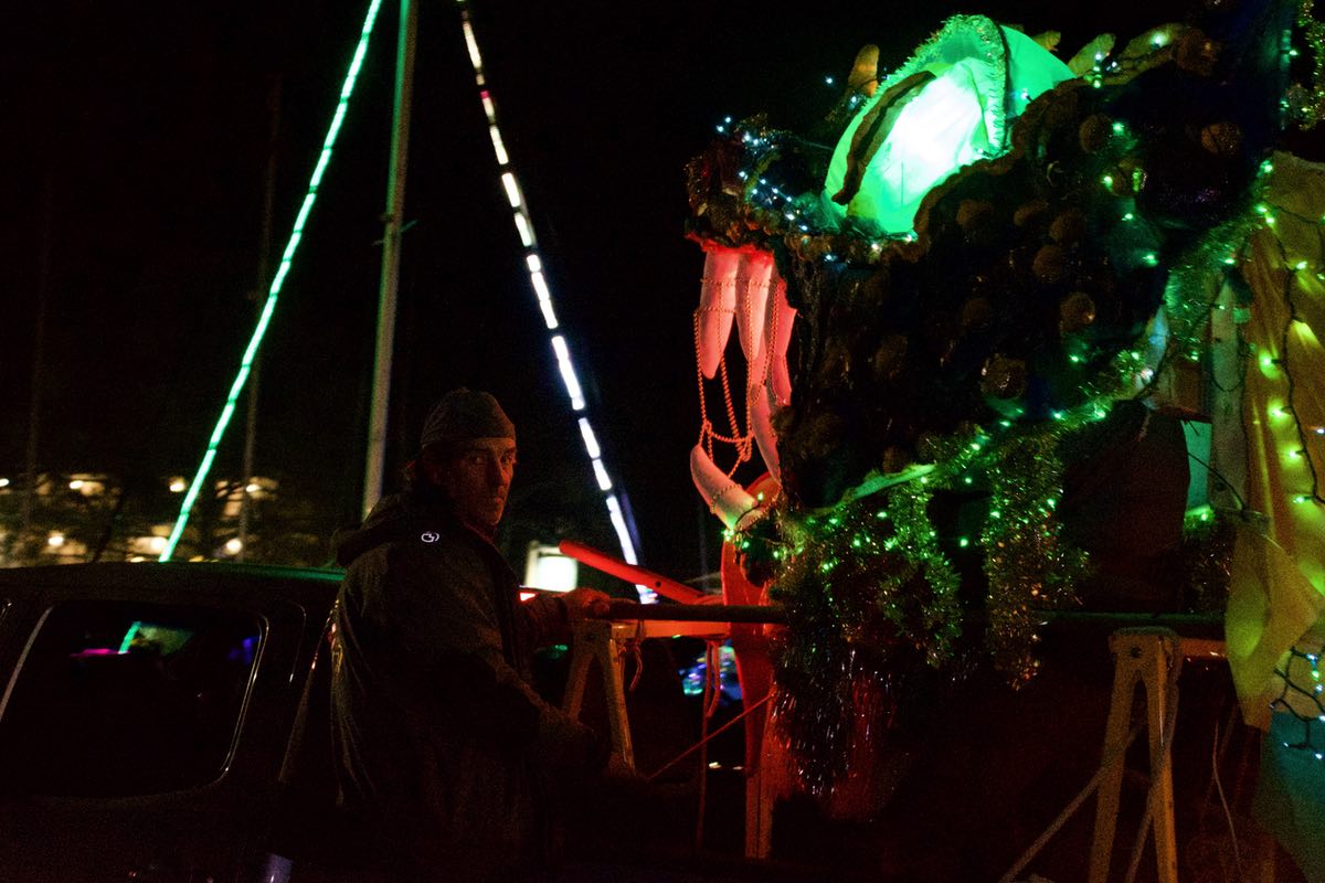 Nighttime. The dragon head has passed by and the green eye is visible. In the background, green lights shine from the mast and mainsail of a boat at the dock. A man with a hat sits on the trailer near the dragon head, looking back at the camera.