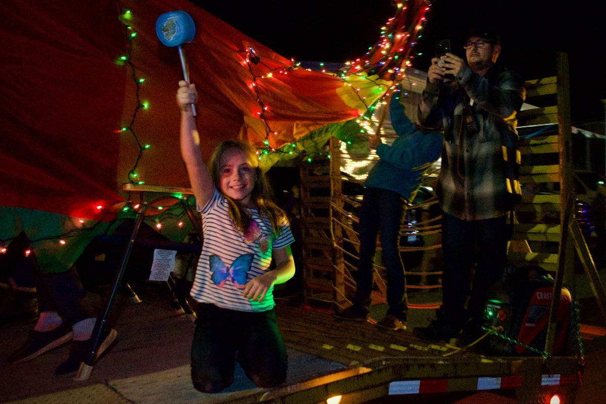A young girl kneels on the trailer beside the dragon's orange, green, and yellow tail, waving a blue noisemaker.