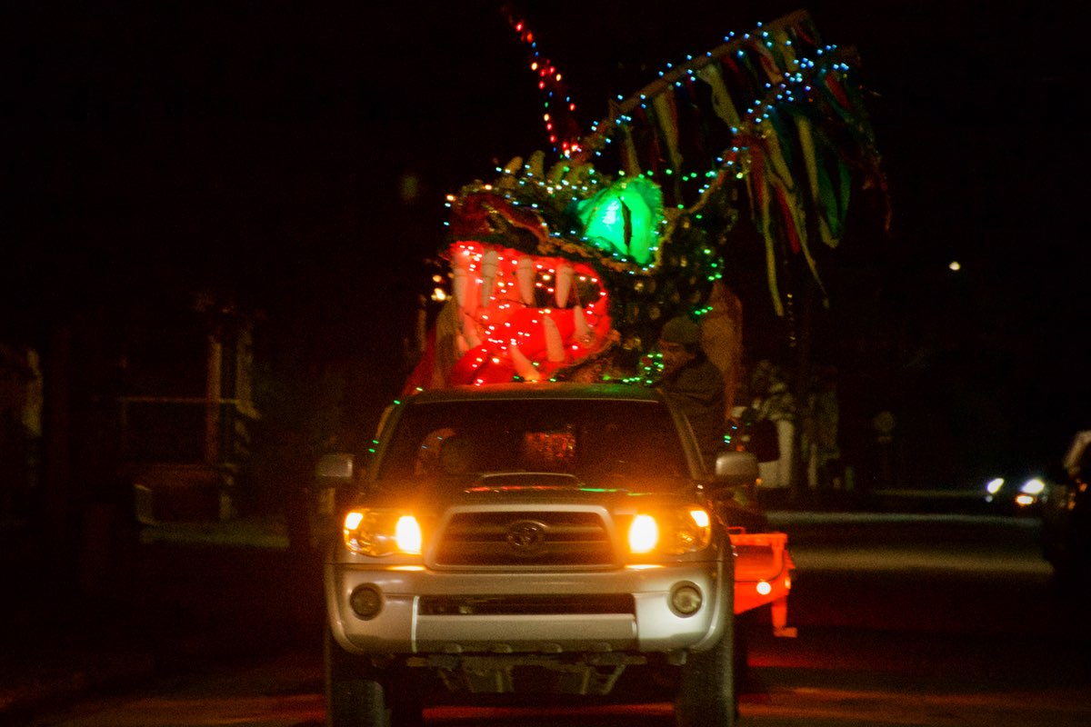 A street at night - a large cloth dragon head draped in colored lights can be seen over the top of pickup truck driving down the road. The dragon has bright green eyes and an open red mouth.